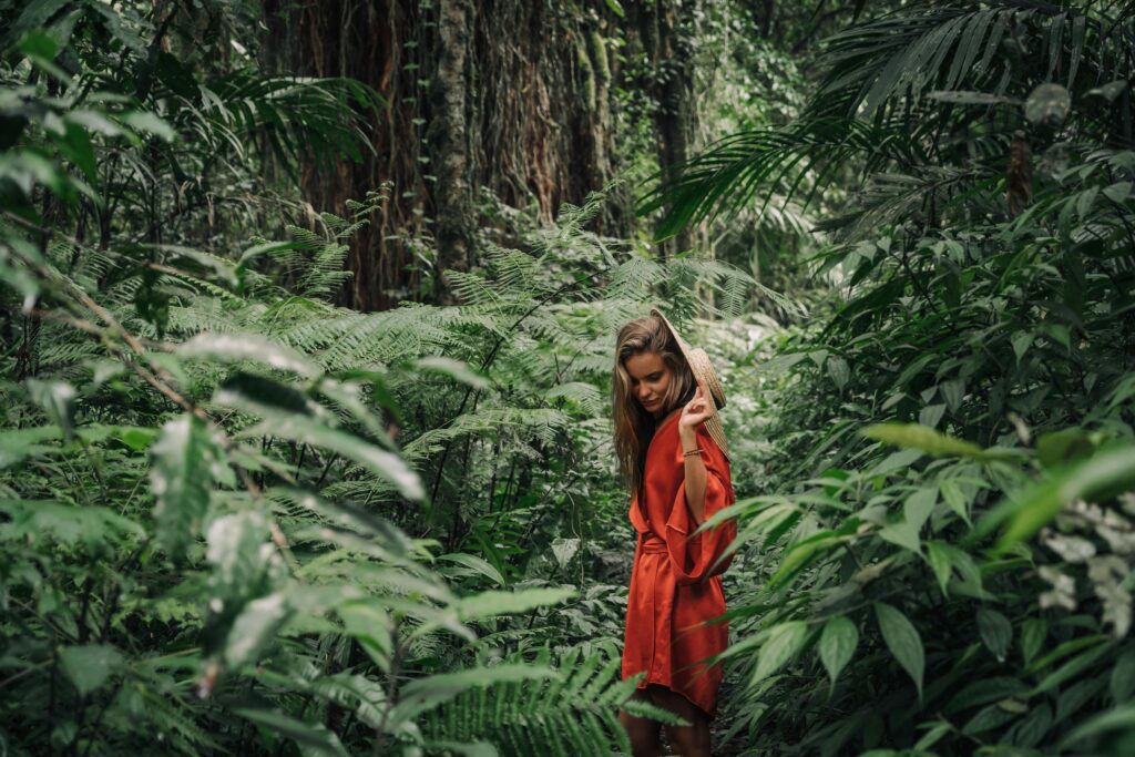A woman stands in a vibrant jungle wearing a red dress, surrounded by lush green foliage.